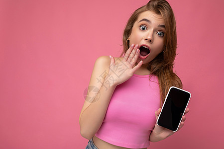 照片中 身穿粉红色上衣的美丽 快乐的金发年轻女子在粉红色背景中与世隔绝 手握空白空间 并展示带有空显示屏的手机 用于模拟看相机互图片