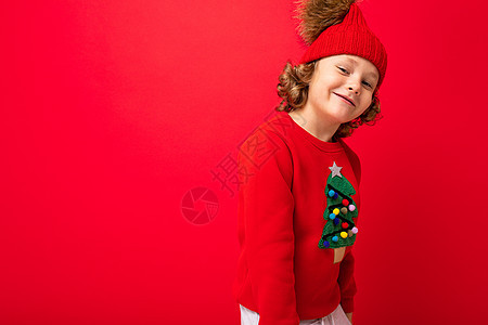 穿着温暖帽子和圣诞毛衣的金发美女男孩 红色背景 面带笑容派对假期微笑男生孩子潮人图片
