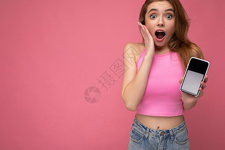 特写照片 令人惊奇的美丽快乐的年轻金发女子穿着粉红色的上衣 在粉红色的背景中与世隔绝 手里拿着空的空间 并展示带有空显示屏的手机图片