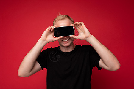 身穿黑色 T 恤 面带微笑 英俊的金发年轻人的肖像 站在红色背景上 与世隔绝 手持手机 手拿智能手机 上面有空显示屏 供模型使用图片