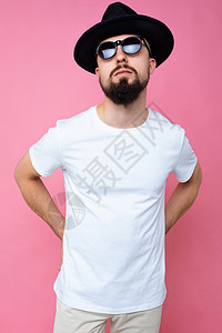 留着胡子的英俊自信的黑发未刮胡子年轻人的垂直照片 他穿着休闲的白色 T 恤 模拟黑色帽子和时尚的太阳镜 在粉红色的背景墙上被隔离图片
