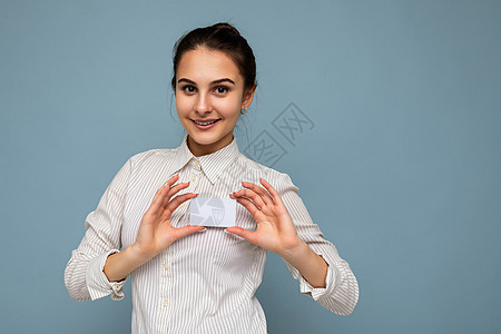 穿着白衬衫的美丽 快乐和笑着微笑的年轻黑发女子 被蓝底持信用卡监视摄影机 隔绝在蓝色背景上图片