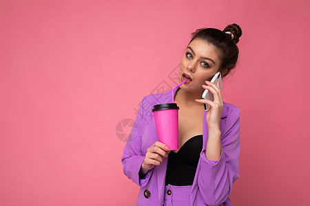 漂亮的黑发女人的肖像穿着潮湿紫色夹克 在智能电话上讲话 用纸杯装着外卖咖啡 与粉红色背景空白广告空间隔绝女士互联网青年饮料西装杯图片