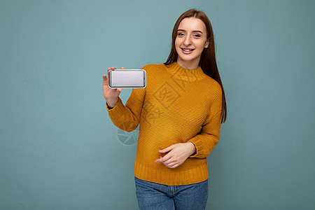 身穿橙色毛衣 面带微笑 积极向上的年轻女性 在蓝色背景中与世隔绝 手握空地 展示手机 留出空地 供模特看着相机图片