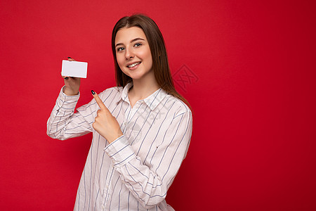照片中 身穿白色衬衫 红色背景中突显的迷人 面带微笑的年轻深色金发女性拿着信用卡 看着相机 手指指着塑料非接触式卡图片