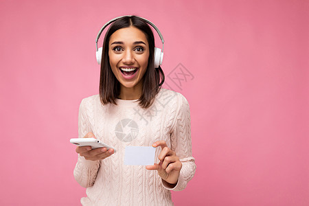 穿着粉色休闲毛衣 戴着白色蓝牙无线耳机 听音乐 使用手机通过信用卡在线支付 看着相机 玩得开心的美丽年轻 微笑的黑发女人的照片 图片