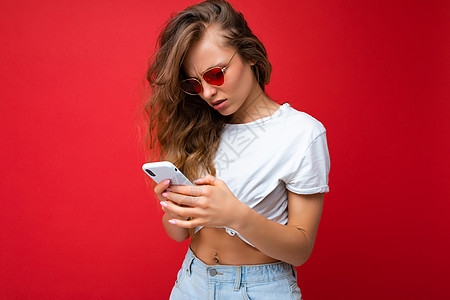 照片中 美丽的年轻女子拿着手机 戴着太阳镜 穿着日常时尚的衣服 隔离在墙壁背景上 看着设备屏幕 在互联网上阅读新闻 不理解图片
