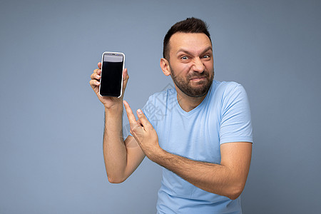 愤怒英俊 未刮胡子的黑发男子穿着日常蓝色 T 恤 与蓝色背景隔离 手持并展示手机 显示屏空空如也 可以看到相机并用手指指着智能手图片