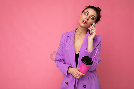漂亮的黑发女人的肖像穿着潮湿紫色夹克 在智能电话上讲话 用纸杯装着外卖咖啡 与粉红色背景空白广告空间隔绝女士杯子女性手机互联网饮图片