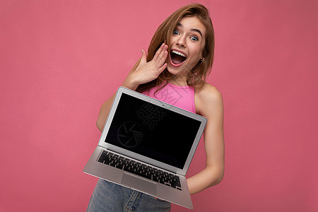 美丽而惊讶的年轻女性手持电脑笔记本电脑 显示器空空如也 头戴粉红色裁剪上衣 看着粉红色背景中突显的相机图片