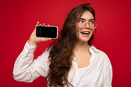 身穿白色衬衫 头戴光学眼镜 身穿红色背景的正面迷人的黑发年轻女性手握手 展示手机 屏幕空空如也 供模特看向侧面图片