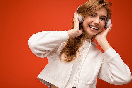 特写照片 迷人 微笑的年轻金发女性穿着白色连帽衫 与彩色背景墙隔开 戴着白色无线蓝牙耳机 听着好听的音乐 玩得开心图片