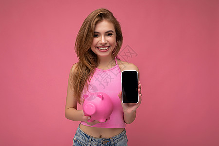 美丽快乐的年轻女性穿着休闲装 与背景隔绝 空空如也 手持手机 展示智能手机 屏幕空空如也 钱箱看起来像粉红猪 看着相机图片