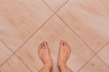 赤脚男子的脚站在浅陶瓷瓷砖地板背景上 从上面看图片