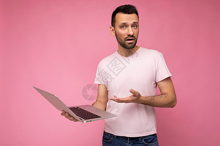 帅气的问黑头发男人拿着笔记本电脑 手看着穿着短袖粉红背景T恤的照相机图片