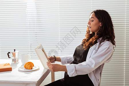 在百叶窗背景下享用早餐 咖啡和羊角面包的快乐年轻漂亮女人看书的侧视图 早上好概念和愉快的午休母性羽绒被女士棕色思维婴儿肚子幸福生图片