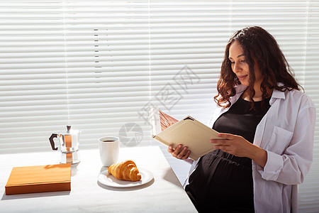 在百叶窗背景下享用早餐 咖啡和羊角面包的快乐年轻漂亮女人看书的侧视图 早上好概念和愉快的午休思维生育羽绒被女士姿势腹部棕色头发商图片