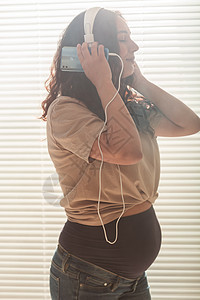 卷发的黑发安抚孕妇使用智能手机和耳机听音乐 在见到婴儿之前舒缓心情的概念腹部母性中年人工作室幸福棕色肚子微笑头发父母图片