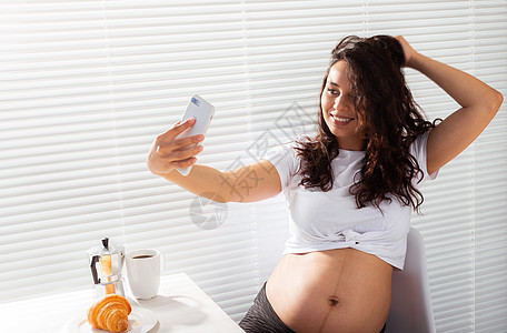 孕妇在早餐时对智能手机采取自拍 产假和技术概念也随之而来 笑声女性生活电话女士照片父母女孩家庭房间婴儿图片