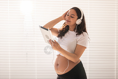 美丽的年轻孕妇通过订阅选择音乐 并在早上通过耳机和平板电脑收听 对胎儿发育有益的概念女士商务腹部生育微笑母性幸福百叶窗黑发羽绒被图片