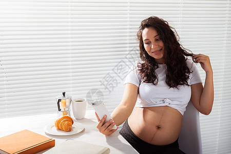 孕妇在早餐时用她的智能手机自画肖像女孕妇摄影腹部闲暇女性快乐技术照片公寓黑发自拍图片