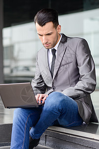 青年商务人士在城市背景的笔记本电脑打字电子产品办公楼人士技术快乐工作套装男性职场管理人员图片
