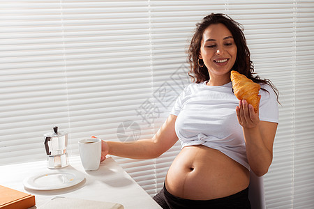 喜悦的怀孕年轻美女在早餐时吃羊角面包和喝茶 上午愉快的早晨概念和与婴儿见面的期望 可复制空间Ropspace女士头发父母姿势微笑图片