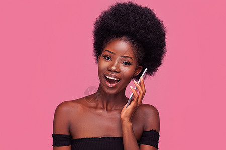 有趣的非洲裔美国女人在电话或智能手机上与女朋友聊天 留着爆炸式发型的女模特在黑色剪裁的上衣中微笑 裸露的肩膀在粉红色背景中突显图片