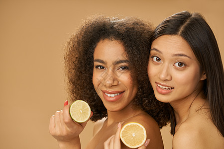 两个可爱的混合种族青年妇女看着相机 把柠檬和石灰切成两半 同时在蜜蜂背景上与众隔绝图片