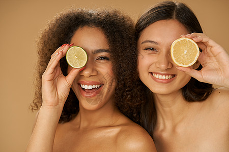 两位快乐的混血年轻女性的肖像看起来很兴奋 用柠檬和酸橙遮住眼睛 切成两半 同时在米色背景中孤立地摆姿势图片
