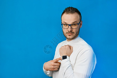 身着白衬衫 眼镜和胡子的年轻有吸引力的欧洲商务人士肖像 手对着蓝色背景的智慧表指着他的长相工人胡须男性导演首席男人人士领带工作成图片