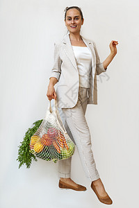 一位身穿浅色西装的女士拿着一个棉质购物袋和装有蔬菜的可重复使用网眼购物袋 没有带复制空间的塑料概念的零浪费 环保净买家图片