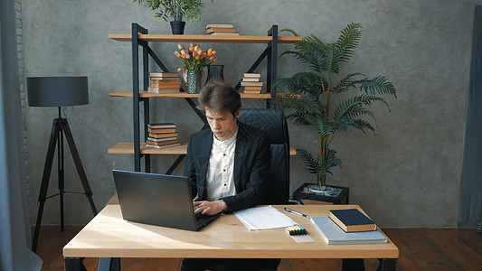 年轻的观点商务人士坐在办公室的桌子旁积极工作 年轻有魅力的商务人士在他办公室的笔记本电脑上工作 年轻英俊的商务人士坐在他的办公室图片