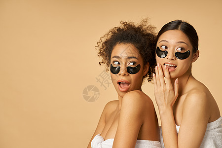 两个玩耍的年轻女性 裹在毛巾上 面部脸部表情令人羡慕 用黑色水凝胶涂在隔着蜜蜂背景的眼罩下图片