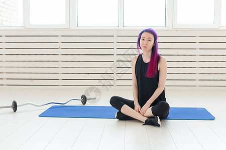 留着紫色头发的年轻混血女嬉皮士坐在杠铃旁边的运动垫上 在健身房里拿着健身球 定期锻炼和健身的概念动机运动装福利运动员护理有氧运动图片