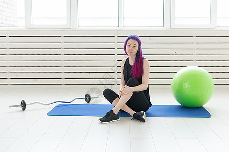 留着紫色头发的年轻混血女嬉皮士坐在杠铃旁边的运动垫上 在健身房里拿着健身球 定期锻炼和健身的概念身体福利有氧运动行动成人活动运动图片