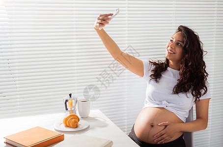 孕妇在早餐时对智能手机采取自拍 产假和技术概念也随之而来 笑声黑发孩子婴儿生活女性父母分娩照片房间肚子图片