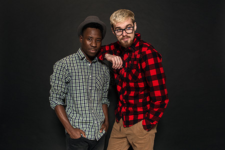 工作室拍摄的两个时尚年轻人玩得开心 穿着衬衫的英俊留着胡子的嬉皮士站在笼子里 站在他的非洲裔美国朋友旁边 戴着帽子 背景是深色的图片