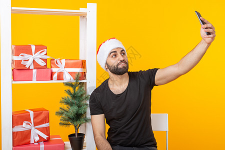 年轻英俊的嬉皮士戴着圣诞老人的帽子 在智能手机上自拍 背景是问候礼物和黄色背景的新年树 新年和圣诞节的概念假期时尚成功人士头发艺背景图片