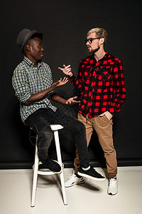工作室拍摄的两个时尚年轻人玩得开心 穿着衬衫的英俊留着胡子的嬉皮士站在笼子里 站在他的非洲裔美国朋友旁边 戴着帽子 背景是深色的图片