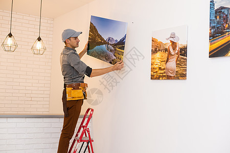 年轻人站在梯子上 而他的妻子帮他在新公寓或房子客厅的墙上挂画图片