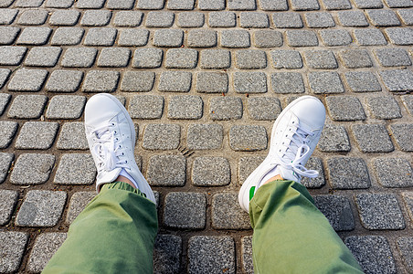 在cobblestone人行道上穿绿色长裤和白色运动鞋的男子腿图片