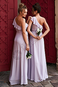 两个漂亮的伴娘女孩 金发女郎和黑发女郎穿着优雅的全长薰衣草紫色薄纱单肩伴娘礼服 手捧鲜花 婚礼当天的欧洲老城区地点图片