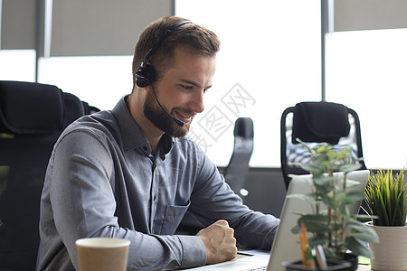 微笑的男性商业顾问戴着耳机坐在现代办公室 看着笔记本电脑屏幕进行视频通话 男士客户服务支持代理帮助热线在线聊天作品中心专业快乐工图片