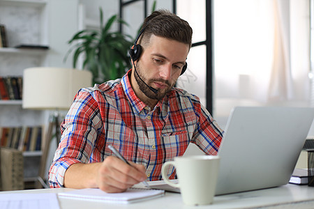 快乐的年轻人用耳机 从家里做笔记本电脑工作 在自我隔离的时候商务微笑男性眼镜学习文书咖啡男人屏幕房子图片