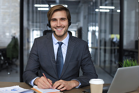 营销中心微笑的男性商业顾问戴着耳机坐在现代办公室 看着笔记本电脑屏幕进行视频通话 男士客户服务支持代理帮助热线在线聊天技术信息销售量软件背景