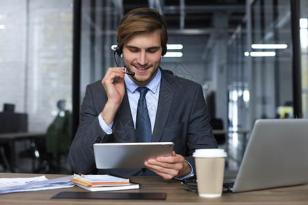 微笑的男性商业顾问戴着耳机坐在现代办公室 看着笔记本电脑屏幕进行视频通话 男士客户服务支持代理帮助热线在线聊天销售量工作者信息联图片