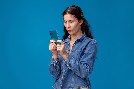 年轻的黑发女人正在装扮着 一个蓝底的智能手机工作室情绪裙子蓝色技术衬衫身体细胞理发女性图片