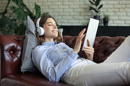 在沙发上用耳机和数字平板电脑微笑的年轻女子享受客厅社交办公笔记本互联网黑发冲浪音乐闲暇图片