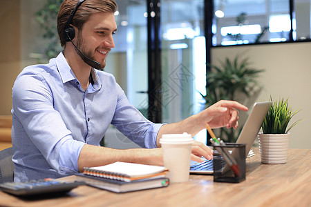 微笑的男性商业顾问戴着耳机坐在现代办公室 看着笔记本电脑屏幕进行视频通话 男士客户服务支持代理帮助热线在线聊天助理销售软件联系远图片